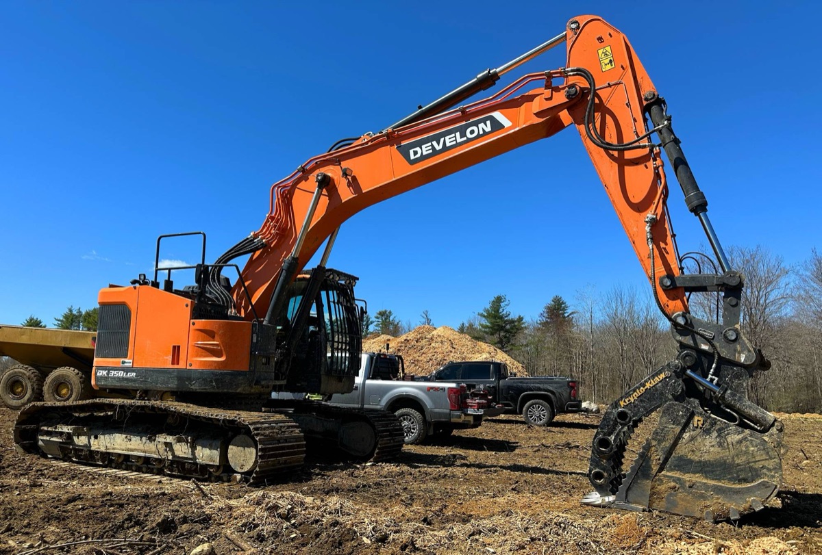 Excavator with a grapple attachment working a cleared job site next to work trucks